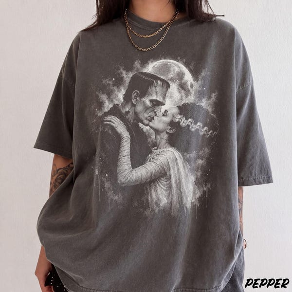 Könnte beinhalten: Ein anthrazitfarbenes Kurzarm-T-Shirt mit einem detaillierten schwarz-weißen Grafikdruck von Frankensteins Monster und seiner Braut, die sich unter einem Vollmond küssen. Das T-Shirt hat einen Rundhalsausschnitt und eine lockere Passform.