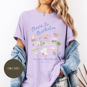 K&ouml;nnte beinhalten: Ein lavendelfarbenes T-Shirt mit einem Grafikdesign, das eine Stadtlandschaft, Palmen und den Text "Cheers to Charleston est. 2023 Lauren's Bachelorette Charleston, SC" zeigt.