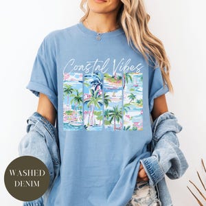 May include: A light blue t-shirt with the text "Coastal Vibes" in white script. The shirt features a graphic print of a tropical scene with palm trees, boats, and buildings.