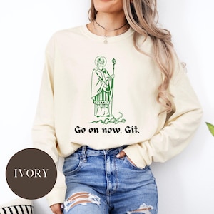 Puede incluir: Una sudadera de manga larga de color crema con una ilustración verde de San Patricio y el texto "Go on now. Git."