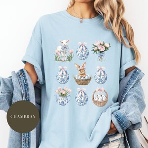 May include: Light blue t-shirt with a pastel Easter theme featuring a lamb, a bunny, floral bouquets, and blue and white eggs. The text "CHAMBRAY" is visible in the lower left corner of the image.