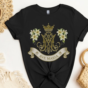 Puede incluir: Camiseta negra con un dise&ntilde;o dorado que presenta una corona, lirios y las letras "Ave Maria".