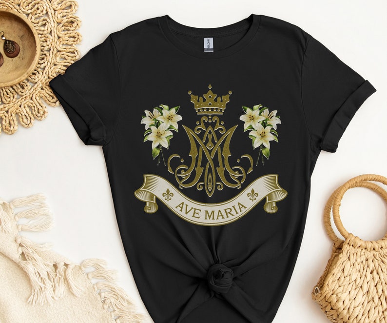 Puede incluir: Camiseta negra con un dise&ntilde;o dorado que presenta una corona, lirios y las letras "Ave Maria".