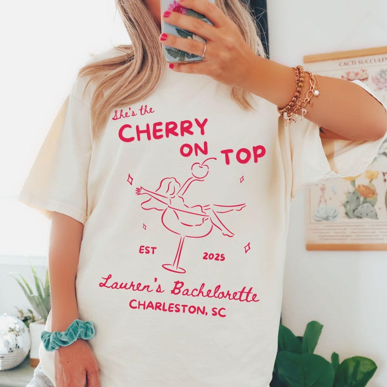 May include: Cream-colored t-shirt with the text "She's the CHERRY ON TOP" in red, along with a graphic of a woman on a cocktail glass. The text "Lauren's Bachelorette, Charleston, SC" is also printed on the shirt. The year "2025" is printed on the shirt.