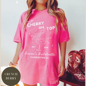 May include: A bright pink t-shirt with the text "He's the Cherry on Top" and "Lauren's Bachelorette Charleston, SC" in white. The shirt features a cocktail graphic and the year 2025. The shirt is a comfortable, oversized fit.