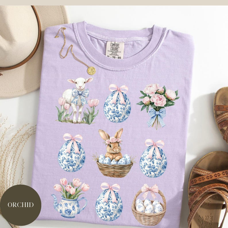 May include: A lavender t-shirt with a pastel Easter design featuring a lamb, a bunny, flowers, and eggs. The eggs are decorated with blue and white floral patterns.