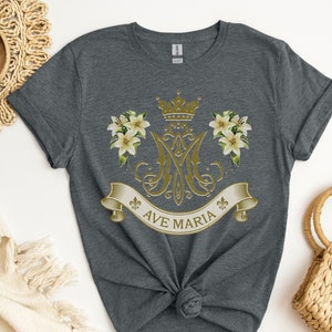 Puede incluir: Una camiseta gris jaspeada con un dise&ntilde;o dorado que presenta una corona, lirios y el texto "AVE MARIA".