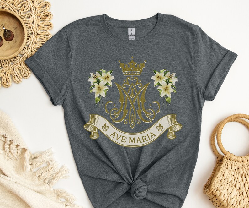 Puede incluir: Una camiseta gris jaspeada con un dise&ntilde;o dorado que presenta una corona, lirios y el texto "AVE MARIA".
