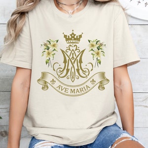 Puede incluir: Una camiseta blanca con un dise&ntilde;o dorado que presenta una corona, lirios y las palabras "Ave Maria".