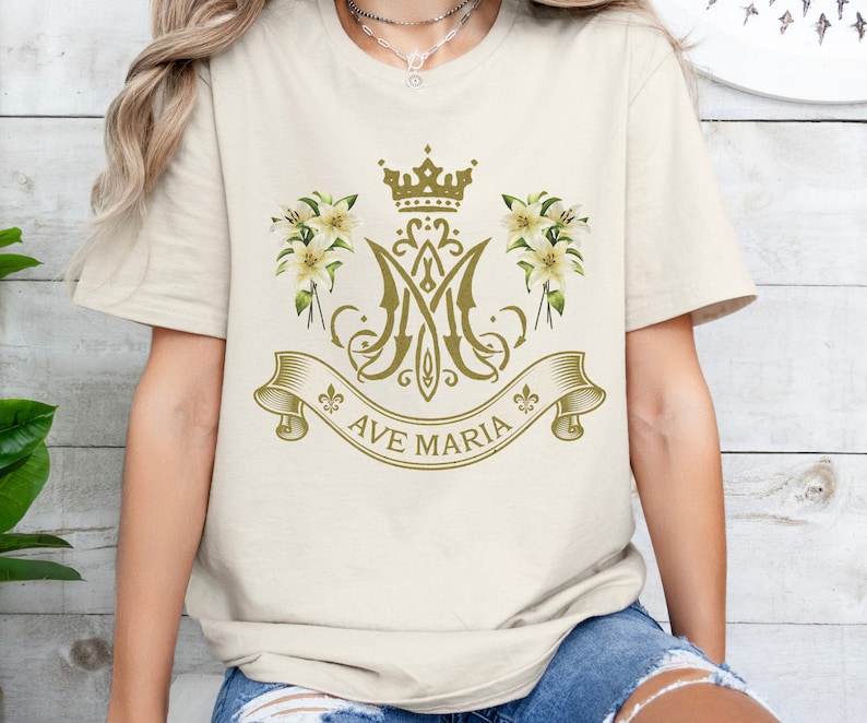 Puede incluir: Una camiseta blanca con un dise&ntilde;o dorado que presenta una corona, lirios y las palabras "Ave Maria".