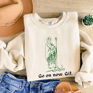 May include: A cream colored sweatshirt with a green illustration of a saint holding a staff. The text "Go on now. Git." is printed below the illustration.