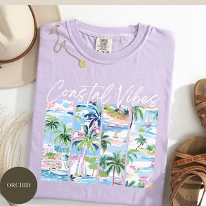 May include: A lavender t-shirt with the text "Coastal Vibes" in white script. The shirt features a graphic of palm trees and sailboats in a tropical scene.