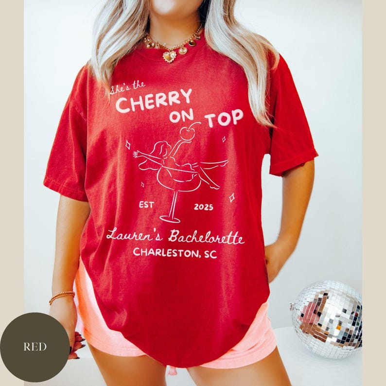 May include: Red t-shirt with white text and a cocktail graphic. The text reads "She's the Cherry on Top" above a cocktail illustration and "Lauren's Bachelorette Charleston, SC". The shirt is a relaxed fit.