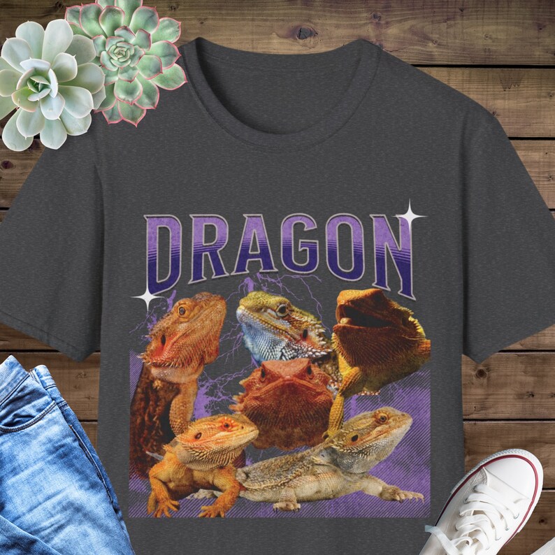 Puede incluir: Una camiseta gris oscuro con un gr&aacute;fico morado y blanco de cinco dragones barbudos. El texto "DRAGON" est&aacute; impreso en letras moradas sobre los dragones.