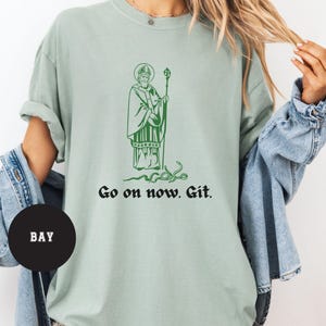 May include: A light green t-shirt with a green illustration of a man in a robe holding a staff. The text "Go on now. Git." is printed below the illustration.