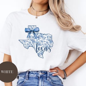 May include: White t-shirt featuring a blue floral Texas outline with a bow and the word "Texas" in script. The shirt is a crew neck and is paired with blue jeans.
