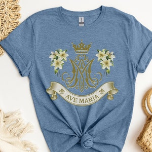 Puede incluir: Una camiseta azul con un dise&ntilde;o dorado de Ave Mar&iacute;a que incluye una corona, lirios y un monograma.
