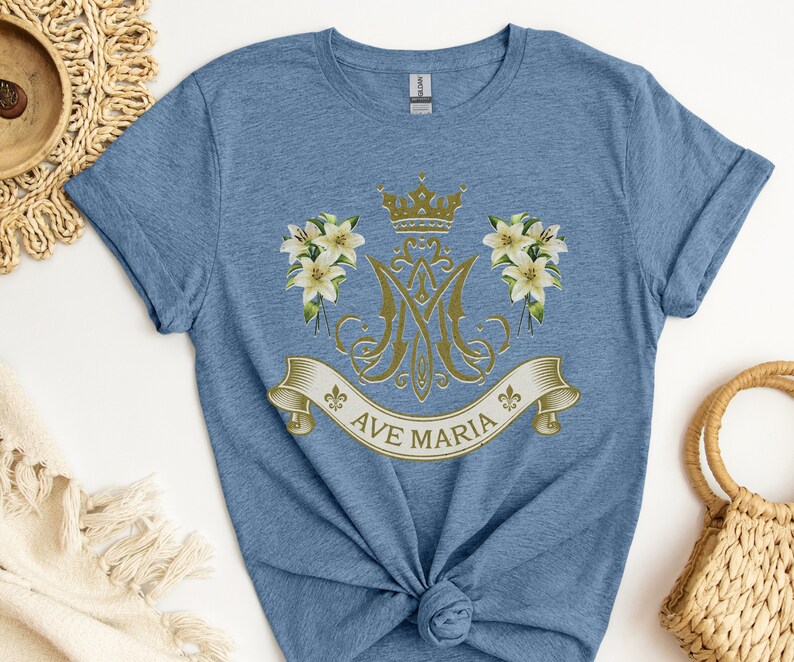 Puede incluir: Una camiseta azul con un dise&ntilde;o dorado de Ave Mar&iacute;a que incluye una corona, lirios y un monograma.