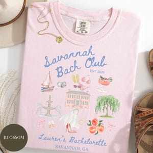 May include: A light pink t-shirt with the words "Savannah Bach Club" and "EST 2026" in blue script. The shirt features illustrations of a sailboat, a house, a fountain, and peaches. The text "Lauren's Bachelorette, Savannah, GA" is also printed on the shirt.