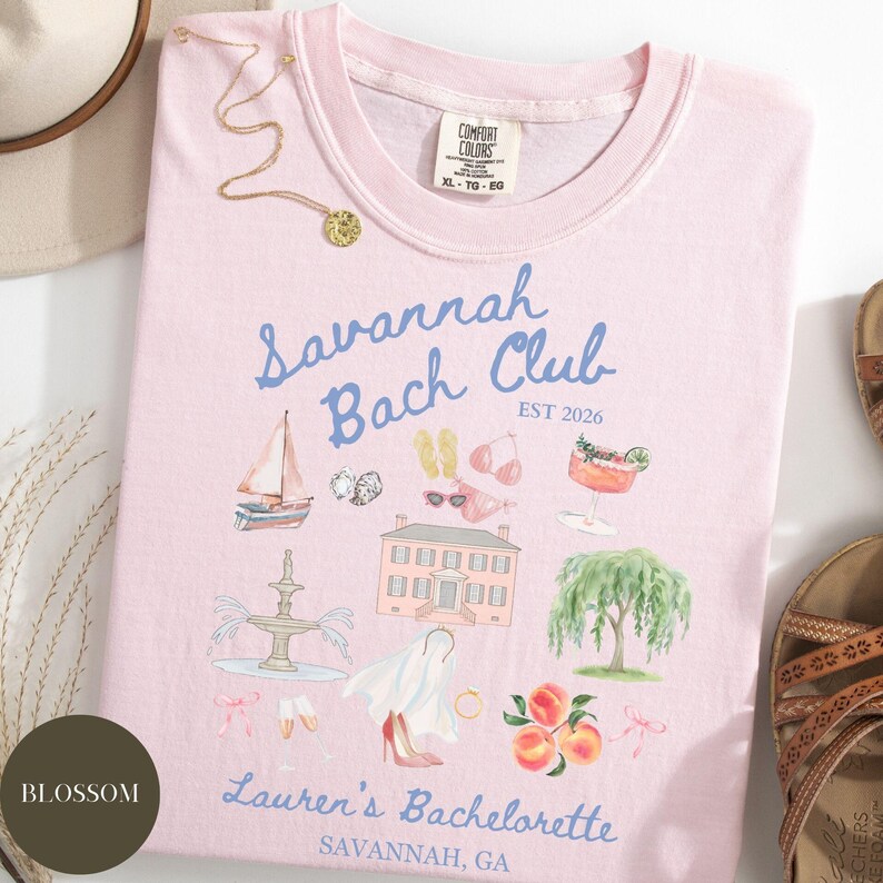 May include: A light pink t-shirt with the words "Savannah Bach Club" and "EST 2026" in blue script. The shirt features illustrations of a sailboat, a house, a fountain, and peaches. The text "Lauren's Bachelorette, Savannah, GA" is also printed on the shirt.