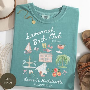 May include: Seafoam green t-shirt with the text "Savannah Bach Club" and illustrations of a sailboat, house, and peaches. The shirt also includes the text "Lauren's Bachelorette" and "Savannah, GA".