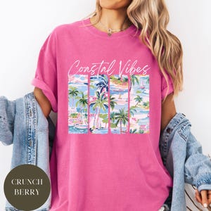 May include: A pink t-shirt with the text "Coastal Vibes" in white lettering. The shirt features a graphic of palm trees and boats in a tropical setting.