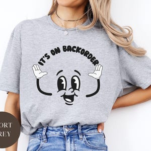 Könnte beinhalten: Ein graues T-Shirt mit dem Text "It's on backorder" in einer Retro-Schrift. Ein Comic-Gesicht mit großen Augen und einem breiten Lächeln wird mit dem Text gezeigt.