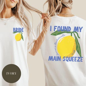 May include: Two white t-shirts with a lemon graphic and text. The first shirt says "Bride" and the second shirt says "I Found My Main Squeeze - Souvenir's Bachelorette Est. 2023".