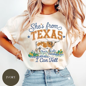 May include: An ivory t-shirt featuring a graphic print with the text "She's from TEXAS I Can Tell" in blue and orange. The design includes a brown longhorn steer standing in a field of green cacti and blue flowers, accented by two small stars.
