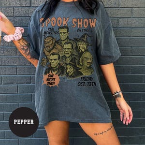 May include: Dark grey t-shirt with a vintage-style graphic featuring classic movie monsters. The text "SPOOK SHOW" is at the top, with "REAL MONSTERS ON STAGE" below. The bottom reads "ONE NIGHT ONLY! FRIDAY OCT. 13TH".