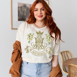 Puede incluir: Una camiseta blanca con un dise&ntilde;o dorado que incluye una corona, detalles florales y el texto "Ave Maria".