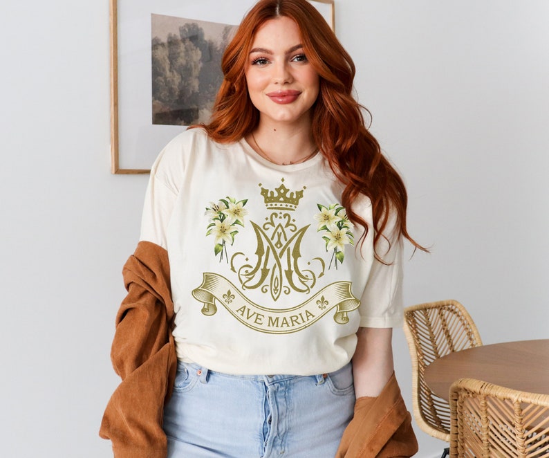 Puede incluir: Una camiseta blanca con un dise&ntilde;o dorado que incluye una corona, detalles florales y el texto "Ave Maria".