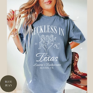 K&ouml;nnte beinhalten: Ein blaues Jeans-T-Shirt mit wei&szlig;em Text, der "Reckless in Texas" lautet, mit einer Grafik von drei Frauen, die ein Martini-Glas halten. Der Text unter der Grafik lautet "Lauren's Bachelorette Austin, TX".