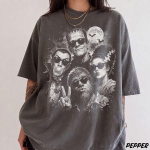 May include: A charcoal gray short-sleeve t-shirt featuring a graphic print of four classic movie monsters wearing sunglasses. The design includes a vampire blowing a bubble, a creature making a peace sign, and a full moon with bats.