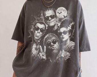 Puede incluir: Una camiseta gris carbón de manga corta con un estampado gráfico de cuatro monstruos de cine clásicos con gafas de sol. El diseño incluye un vampiro haciendo una pompa de chicle, una criatura haciendo el signo de la paz y una luna llena.
