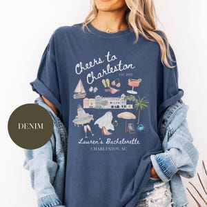 K&ouml;nnte beinhalten: Ein marineblaues T-Shirt mit einem wei&szlig;en Grafikdesign, auf dem "Cheers to Charleston Est. 2025" steht, mit Illustrationen eines Segelboots, einer Sonnenbrille, Flip-Flops, eines Cocktails, Geb&auml;uden, einer Braut, eines Rings, eines Schirms, eines Strandstuhls und einer Krabbe. Der Text "Lauren's Bachelorette Charleston, SC" ist ebenfalls auf dem Shirt enthalten.