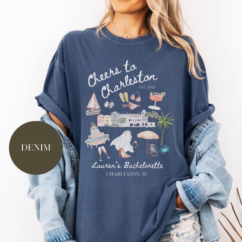 K&ouml;nnte beinhalten: Ein marineblaues T-Shirt mit einem wei&szlig;en Grafikdesign, auf dem "Cheers to Charleston Est. 2025" steht, mit Illustrationen eines Segelboots, einer Sonnenbrille, Flip-Flops, eines Cocktails, Geb&auml;uden, einer Braut, eines Rings, eines Schirms, eines Strandstuhls und einer Krabbe. Der Text "Lauren's Bachelorette Charleston, SC" ist ebenfalls auf dem Shirt enthalten.