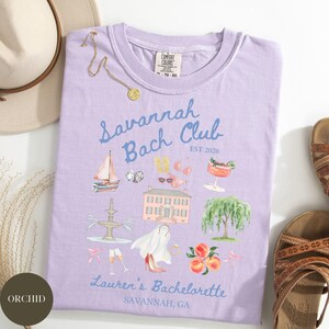 May include: A light purple t-shirt with the text "Savannah Bach Club" and illustrations of a sailboat, house, and peaches. The shirt also says "Lauren's Bachelorette, Savannah, GA". A gold necklace and sandals are also visible.