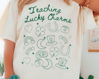Puede incluir: Una camiseta blanca con el texto "Teaching Lucky Charms" en escritura verde. La camiseta está decorada con contornos verdes de amuletos de la suerte, incluyendo corazones, tréboles, herraduras, arcoíris, estrellas, lunas y ollas de oro.