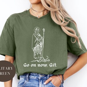 May include: A military green t-shirt with a white graphic of a saint holding a staff and a snake. The text "Go on now. Git." is printed below the graphic.