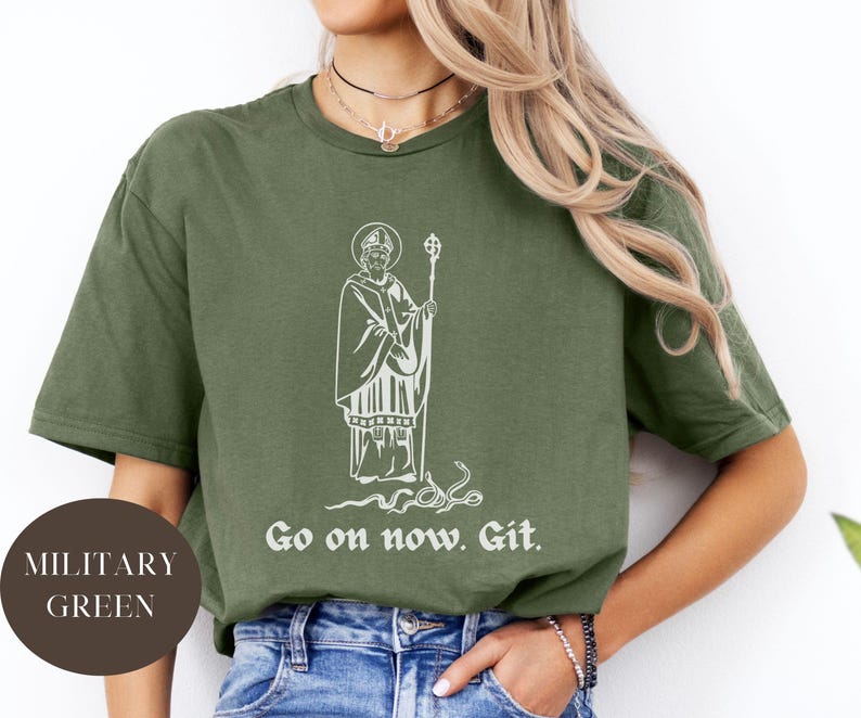 May include: A military green t-shirt with a white graphic of a saint holding a staff and a snake. The text "Go on now. Git." is printed below the graphic.