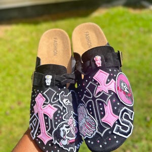 Könnte beinhalten: Schwarze Clogs mit rosa Kreuz- und Totenkopf-Patches, verziert mit silbernen Nieten. Die Clogs haben einen schwarzen Riemen und ein Korkfußbett. Das Wort "FASHION" ist auf dem Fußbett aufgedruckt.