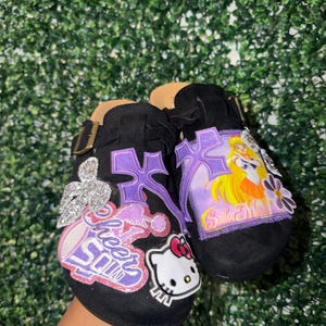Könnte beinhalten: Ein Paar schwarze Schlupfschuhe mit dekorativen Aufnähern, darunter eine Sailor Moon-Figur, ein Hello Kitty-Gesicht, eine glitzernde silberne Blume und ein violettes Kreuz. Die Schuhe haben einen Riemen mit goldfarbener Schnalle und ein beiges Fußbett.