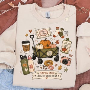 May include: A beige long-sleeve sweatshirt featuring a graphic print of a green wagon filled with pumpkins, a coffee cup, a snack pouch, a camera, hair ties, and socks. The text reads "PUMPKIN PATCH LOGISTICS DEPARTMENT" with small decorative icons.