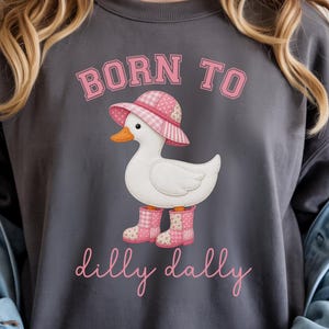 Op de afbeelding: Donkergrijze sweatshirt met een afbeelding van een witte eend met een roze hoed en laarzen. De tekst "BORN TO" en "dilly dally" staat in roze letters. De eend heeft een oranje snavel.