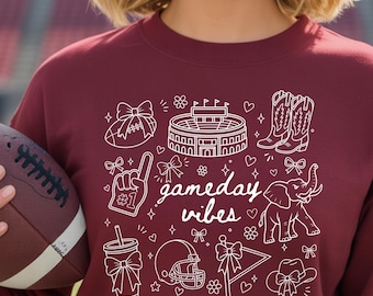 Può includere: Una felpa bordeaux a maniche lunghe con una grafica a linee bianche che riporta il testo gameday vibes. Il design include illustrazioni di un pallone da football, uno stadio, stivali da cowboy, un dito di gommapiuma, un elefante e un casco.