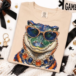 May include: A beige crew neck t-shirt featuring a graphic print of an alligator wearing orange-rimmed sunglasses and a chunky necklace with large blue and orange rectangular gemstones. The shirt has a Comfort Colors brand label at the inner neckline.