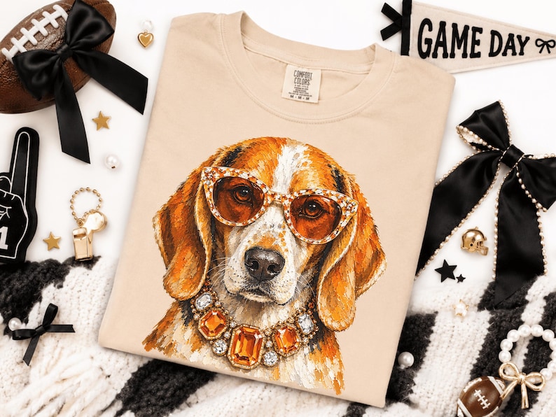 May include: A beige t-shirt featuring a graphic illustration of a beagle dog wearing orange-tinted cat-eye sunglasses and a large, ornate necklace made of amber-colored and clear crystals. The shirt has a Comfort Colors brand label at the neck.