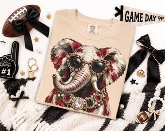 May include: A beige t-shirt featuring a graphic of an elephant wearing jeweled sunglasses and a large, ornate gemstone necklace. The elephant is rendered in shades of red, white, and black. The shirt has a Comfort Colors brand tag at the neck.