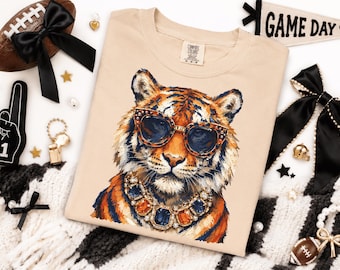 May include: A beige t-shirt featuring a graphic illustration of a tiger wearing leopard-print sunglasses and a chunky, multi-colored gemstone necklace. The tiger is depicted with orange, black, and white fur, and the shirt has a Comfort Colors label.